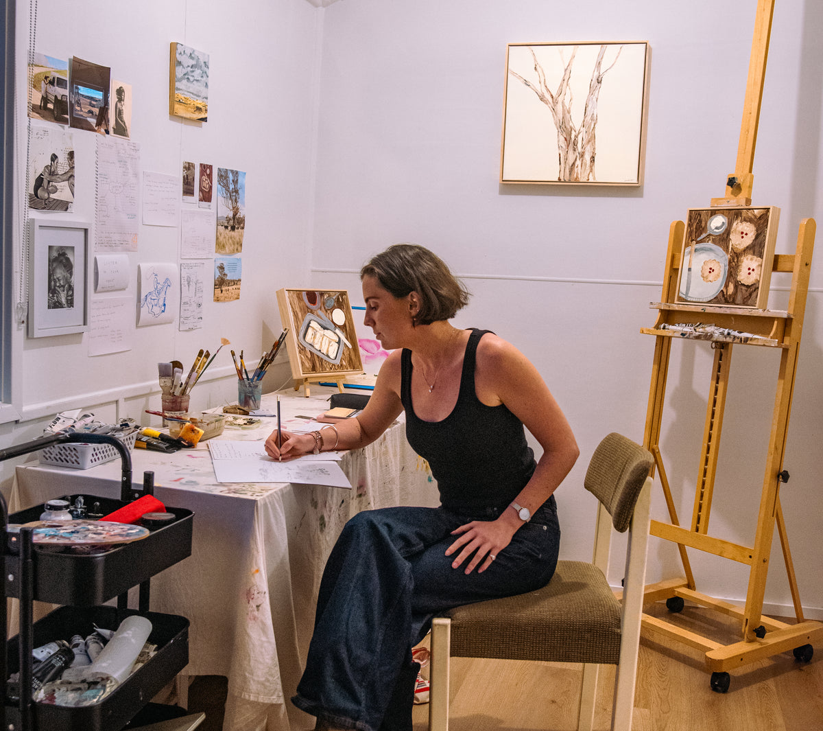 A woman in a black tank top and dark jeans sits at a small artist's desk, writing on paper. The room has white walls decorated with art prints and sketches, a wooden easel with a painting, and an artist's cart with supplies.